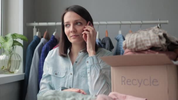 Seorang wanita mengumpulkan pakaian dalam kotak untuk daur ulang atau pembuangan. seorang wanita Millennial panggilan layanan daur ulang dari smartphone nya. — Stok Video