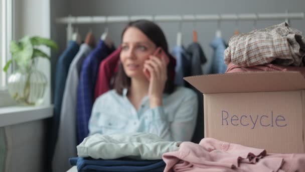 Een vrouw verzamelt kleding in een doos voor recycling of verwijdering. een duizendjarige vrouw belt de recyclingdienst vanaf haar smartphone. — Stockvideo