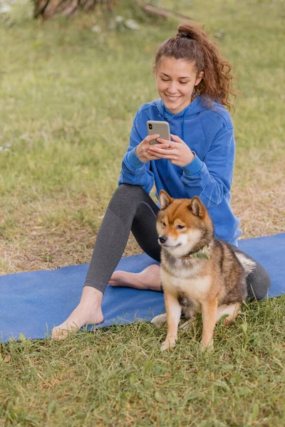 Mujer con un perro en el parque hace deporte o fitness en una esterilla de yoga. una mujer europea con el pelo rizado en una sudadera con capucha azul está charlando en un teléfono inteligente — Foto de Stock