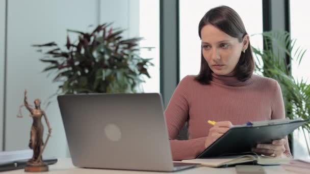 Een vrouwelijke advocaat in haar kantoor werkt aan een laptop en maakt aantekeningen in een notitieboekje. een Europese notaris typt op een laptop. — Stockvideo