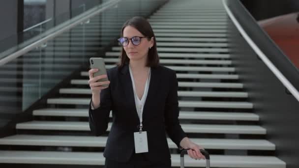 Geschäftsfrau im Businessanzug mit Koffer läuft durch den Flughafen oder das Business Center und telefoniert. eine selbstbewusste Europäerin — Stockvideo
