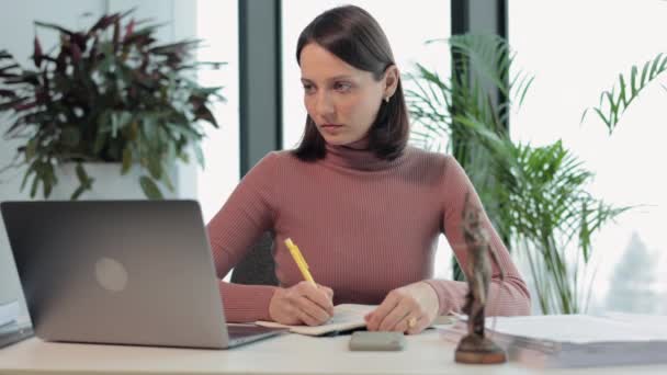 Un avvocato donna nel suo ufficio sta lavorando su un computer portatile e prendere appunti in un quaderno. una notaia europea sta digitando su un computer portatile. — Video Stock