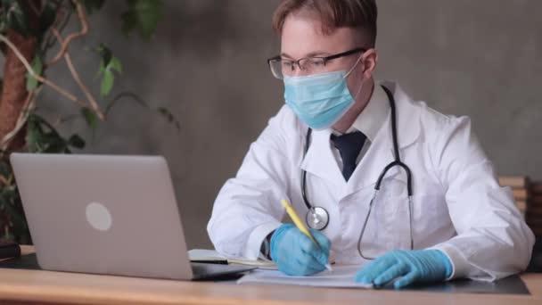 Beyaz önlüklü, tıbbi maskeli ve eldivenli erkek doktor, bir dizüstü bilgisayardaki webcam ile bir hasta randevusu düzenler ve bir not defteri veya reçete yazar — Stok video