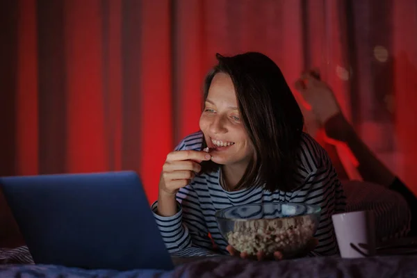 Frau in gestreiftem Sweatshirt und Hut isst Popcorn und schaut zu Hause vom Laptop aus einen Film im Fernsehen oder eine Fernsehserie — Stockfoto