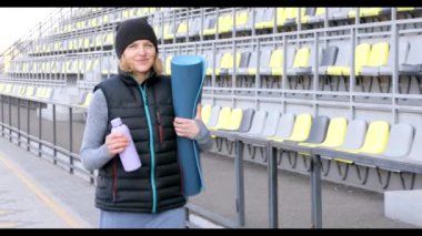 Genç ve çekici bir kadın sabah spor yapıyor. Parkta güneş doğuyor. Elinde yoga minderi ve su şişesi var.