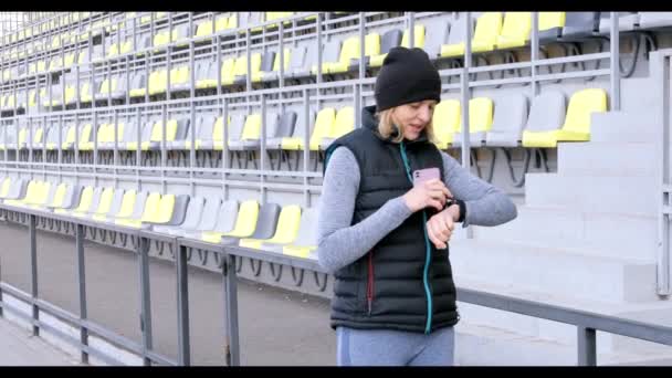 Mulher com telefone celular conectado a um relógio inteligente. Desportista olhando para smartwatch e segurando telefone inteligente em sua outra mão, ao ar livre — Vídeo de Stock