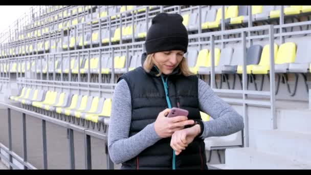Žena s mobilním telefonem připojená k chytrým hodinkám. Sportswoman dívá na Smartwatch a drží chytrý telefon v druhé ruce, venku — Stock video