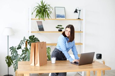 Ev ofisinde çalışan mutlu iş kadını hediye çantaları ve ambalajlarla çevrili dizüstü bilgisayarına bakıyor.