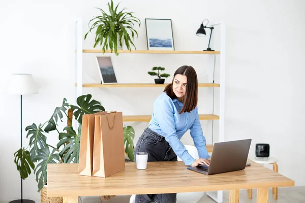Ev ofisinde çalışan mutlu iş kadını hediye çantaları ve ambalajlarla çevrili dizüstü bilgisayarına bakıyor.