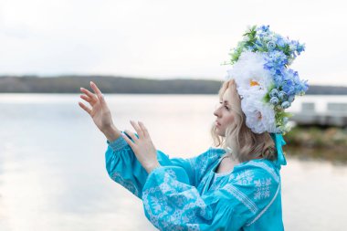 Güzel, genç Ukraynalı, mavi işlemeli bir elbise giymiş, başında büyük güzel bir çiçek çelengi olan bir kadın ellerini gölün manzarasının önünde uzatıyor..