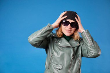 Siyah şapkalı neşeli kız ellerini şakacı bir şekilde kafasında tutuyor, eğlenceli gülüyor, yeşil ceket giyiyor.