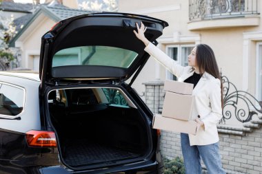 Young business woman picking up parcels from a car trunk, coming home by car. The girl presses a button to close the trunk.