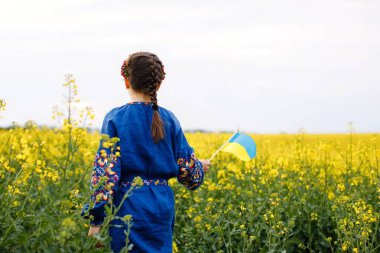 Ukrayna için dua et. Kolza tarlasında Ukrayna bayrağı taşıyan bir çocuk. Nakışlı gömlekli bir kız elinde Ukrayna bayrağıyla sahada koşuyor.
