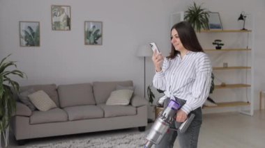 Mutlu, genç, beyaz, güzel kadın yerleri süpürüyor ve evde akıllı telefon kullanıyor.