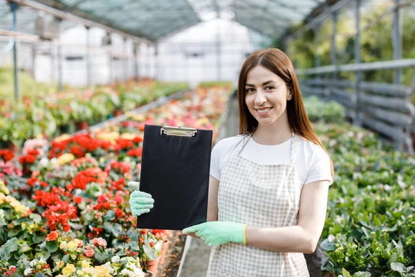Portrait of gardener woman at work in greenhouse with notebook examines the growing flowers in greenhouse.