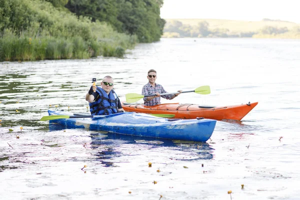 Kayaking on the river. Two young men sit in kayaks and take a selfie with a portable camera. The concept of water entertainment.