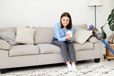 Büyüleyici esmer ev hanımı modern süpürgeyle halıyı temizledikten sonra evde eğleniyor.