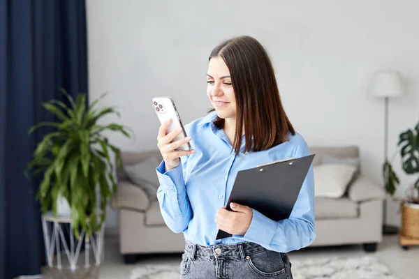 Young woman chatting by the phone and smiling, standing on the middle of room, got message from family, talking by video, long-distance relationship