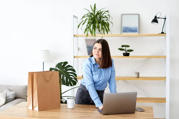 Happy businesswoman working in a home office standing looking at her laptop computer surrounded by gift bags and packaging