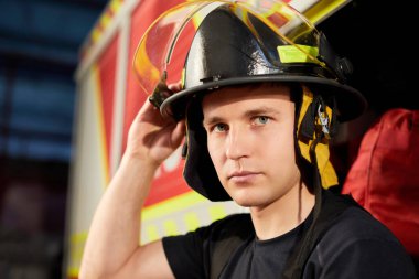 Kaslı, yakışıklı bir itfaiyecinin portresi itfaiye arabasının arka planında, elinde yangın kaskı var.