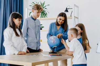 Teacher and children in class are looking at globe, teacher helps explain the lesson to the children in the class at a desk. Educational school process, bright room and interesting learning