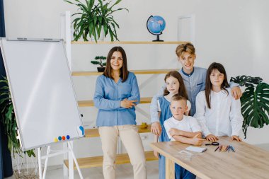 Birlikte gülümsüyoruz. Farklı yaşlarda bir grup çocuk okulda öğretmenle birlikte geniş bir sınıfta duruyor..