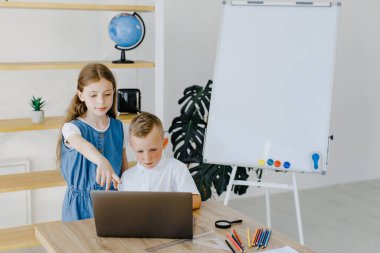 Ergen kız ve birinci sınıf öğrencisi çocuk laptopta birlikte ders çalışıyorlar sınıfta masa başında oturuyorlar. Genç kız ve birinci sınıf çocuğu okulda bilgisayar projesinde beraberler.