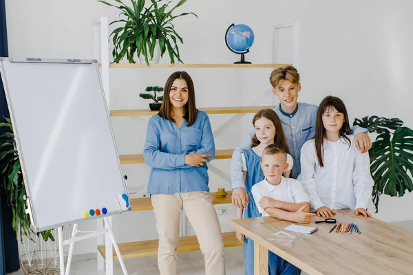 Birlikte gülümsüyoruz. Farklı yaşlarda bir grup çocuk okulda öğretmenle birlikte geniş bir sınıfta duruyor..