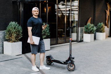 Caddedeki elektrikli scooter 'da dizüstü bilgisayarı ve telefonu elinde tutan yakışıklı bir serbest çalışan.