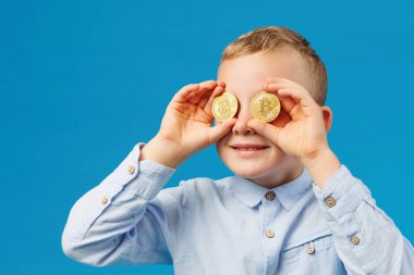 Kripto para ve modern finans. Neşeli çocuk stüdyoda mavi arka planda altın Bitcoin tutuyor ve gözlerini bozuk paralarla kapatıyor.