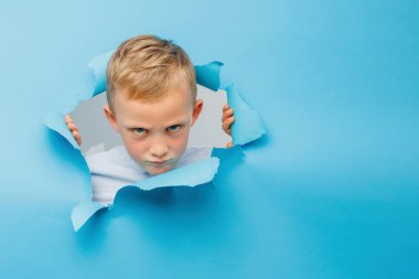 Mutlu şirin çocuk mavi arka plan duvarında eğleniyor, kağıttaki bir delikten tırmanıyor. Çocuğun neşeli ve komik duyguları.