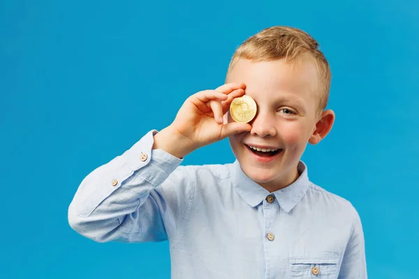 Cryptocurrency and modern finance. Cheerful boy holding golden bitcoin in studio on the blue background and covers his eye with a coin