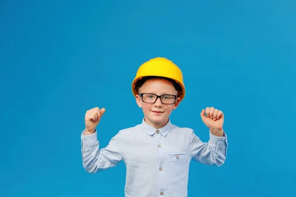 Cute boy construction worker in a yellow hard hat stands in an indoor studio on a blue background and shows satisfaction with the work done. Future construction worker