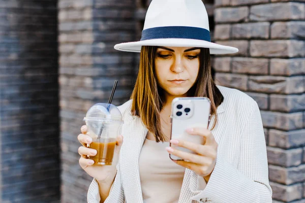 Beautiful woman going to meeting with cold coffee and holding the phone walking on the street. Portrait of successful business woman holding cup of cold drink in hand on her way to work on city street