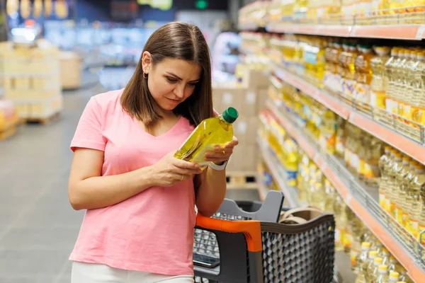 Young pretty girl is shopping in a big store. The girl buys groceries at the supermarket. Beautiful smiling young woman in casual clothes is holding two bottles of olive oil in the supermarket.