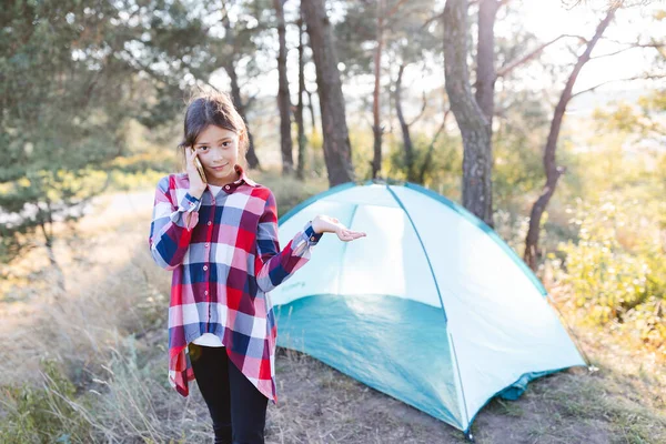 The girl talking on a mobile phone. Teenager girl standing near a tent and holding mobile phone in hand. Remote communication. Camping in nature.