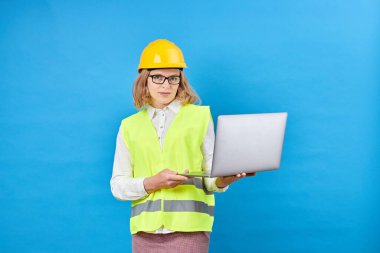Profesyonel kadın mühendislik ustabaşının stüdyo görüntüleri sert kask ve yansıtıcı güvenlik yeleği giyer, ayakta durur ve gülümser, dizüstü bilgisayarı mavi arka planda tutar.