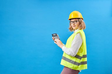 Profesyonel kadın mühendislik ustabaşının stüdyo görüntüleri sert kask ve yansıtıcı güvenlik yeleği giyer, ayakta durur ve gülümser, hesap makinesini mavi arka planda tutar.