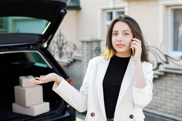 若い実業家の女性が小包を車で帰宅して車の中で立っている 女の子は立って車の近くの携帯電話で話している — ストック写真