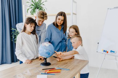 Teacher and children in class are looking at globe, teacher helps explain the lesson to the children in the class at a desk. Educational school process, bright room and interesting learning