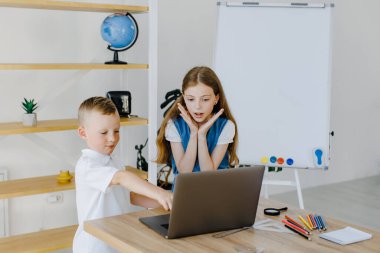 Teenage girl and first grader boy study on laptop together sitting at desk in classroom. Teenage girl and first grader boy do project on computer together at school