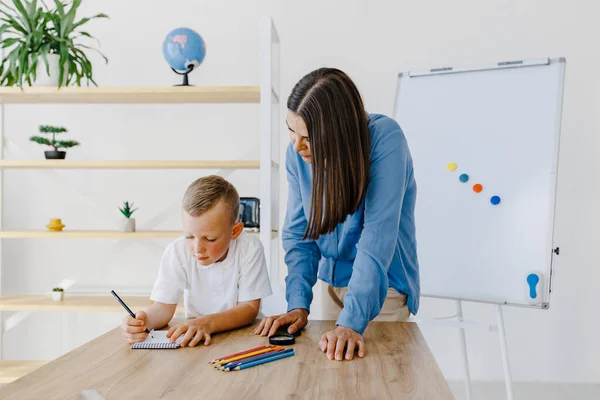 Private lesson. Attentive young woman tutor teacher helping little boy pupil with studying, correct mistakes explain learning material. Smiling mother assist small boy with home task