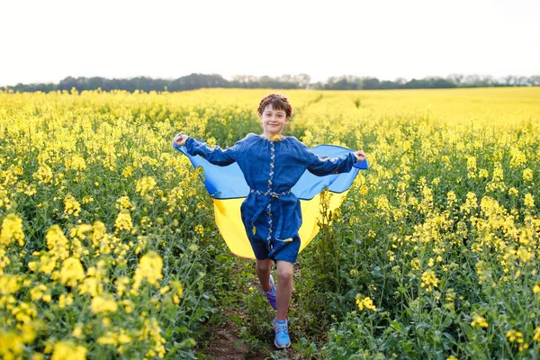 Pray for Ukraine. Child with Ukrainian flag in rapeseed field. A girl in an embroidered shirt runs across the field with the Ukrainian flag in her hands