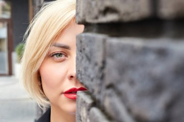 Beyaz bir dış duvara yaslanmış, kenar mahallede fotokopi alanı olan ve kameraya bakan sevimli genç bir kadın.