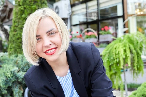 Blonde woman dressed in business style sits on a chair and looks at the camera