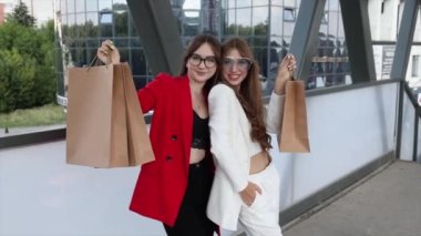Two happy women dress in stylish suits are standing together in front of the shopping mall with a lots of paper bags.