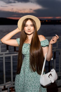 Portrait romantic young woman dressed in green dress and hat near little lake. Girl walks at the park in evening.