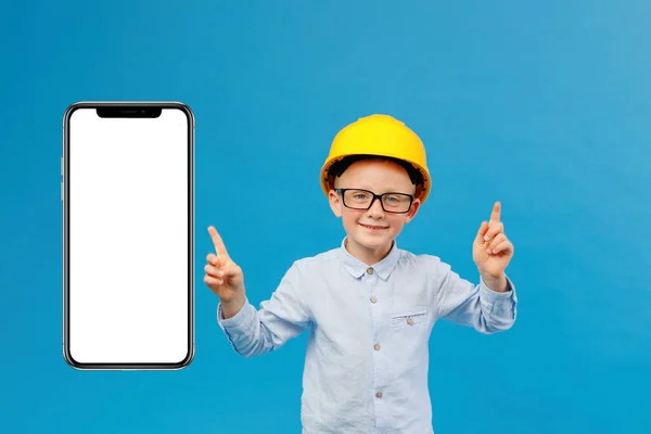 Cute boy construction worker in yellow hard hat standing in indoor studio on blue background and pointing direction with fingers near huge mobile phone. Future construction worker