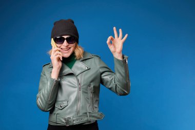 Cheerful pretty woman in bright green jacket holds smartphone in her hand and showing OK sign, gesturing with fingers, smiling cutely looking at camera, isolated over blue background 