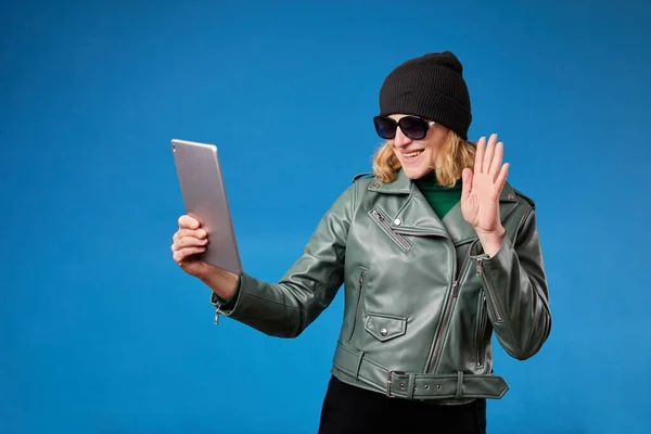 Studio shot of young female model in green jacket standing smiling holding tablet computer video call waving hand say hi hello greeting goodbye